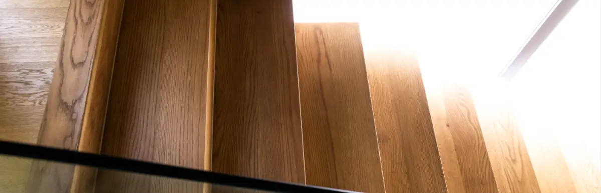 Eine Nahaufnahme aus einem hohen Blickwinkel zeigt eine moderne Holztreppe mit Glasgeländer. Die Treppe besteht aus hellbraunem Holz mit sichtbarer Maserung und führt von links unten nach rechts oben im Bild. Ein elegantes Glasgeländer mit einem dunklen Metallrahmen verläuft entlang der linken Seite der Treppe und ist teilweise im Vordergrund zu sehen. Der obere Teil der Treppe ist in helles, natürliches Licht getaucht, was ein Gefühl von Offenheit und Wärme vermittelt. Die Komposition ist klar und minimalistisch und betont die natürliche Schönheit des Holzes und das architektonische Design der Treppe.