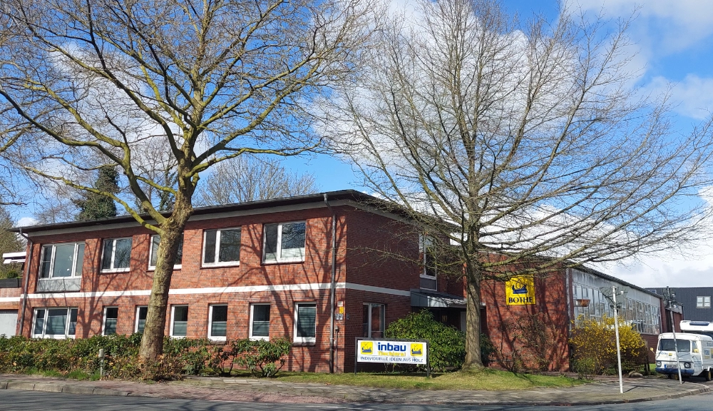 Eine Weitwinkelaufnahme aus niedriger Perspektive zeigt ein zweistöckiges Backsteingebäude mit Flachdach unter strahlend blauem Himmel mit vereinzelten weißen Wolken. Zwei große, kahlastige Bäume dominieren den Vordergrund, ihre Äste ragen über den Bildausschnitt hinaus und verdecken teilweise das Gebäude. Das Gebäude hat zahlreiche Fenster mit weißen Rahmen, und zwischen dem ersten und zweiten Stock verläuft horizontal ein weißer Streifen. An der Vorderseite des Gebäudes ist ein Schild mit gelber und blauer Schrift mit der Aufschrift „inbau” zu sehen, zusammen mit einem kleineren Schild mit der gelben Aufschrift „BOHHE”. Auf der rechten Seite des Bildes, in der Nähe des Eingangs des Gebäudes, steht ein weißer Lieferwagen. Der Boden vor dem Gebäude ist mit grünem Gras und niedrigen Büschen bewachsen.